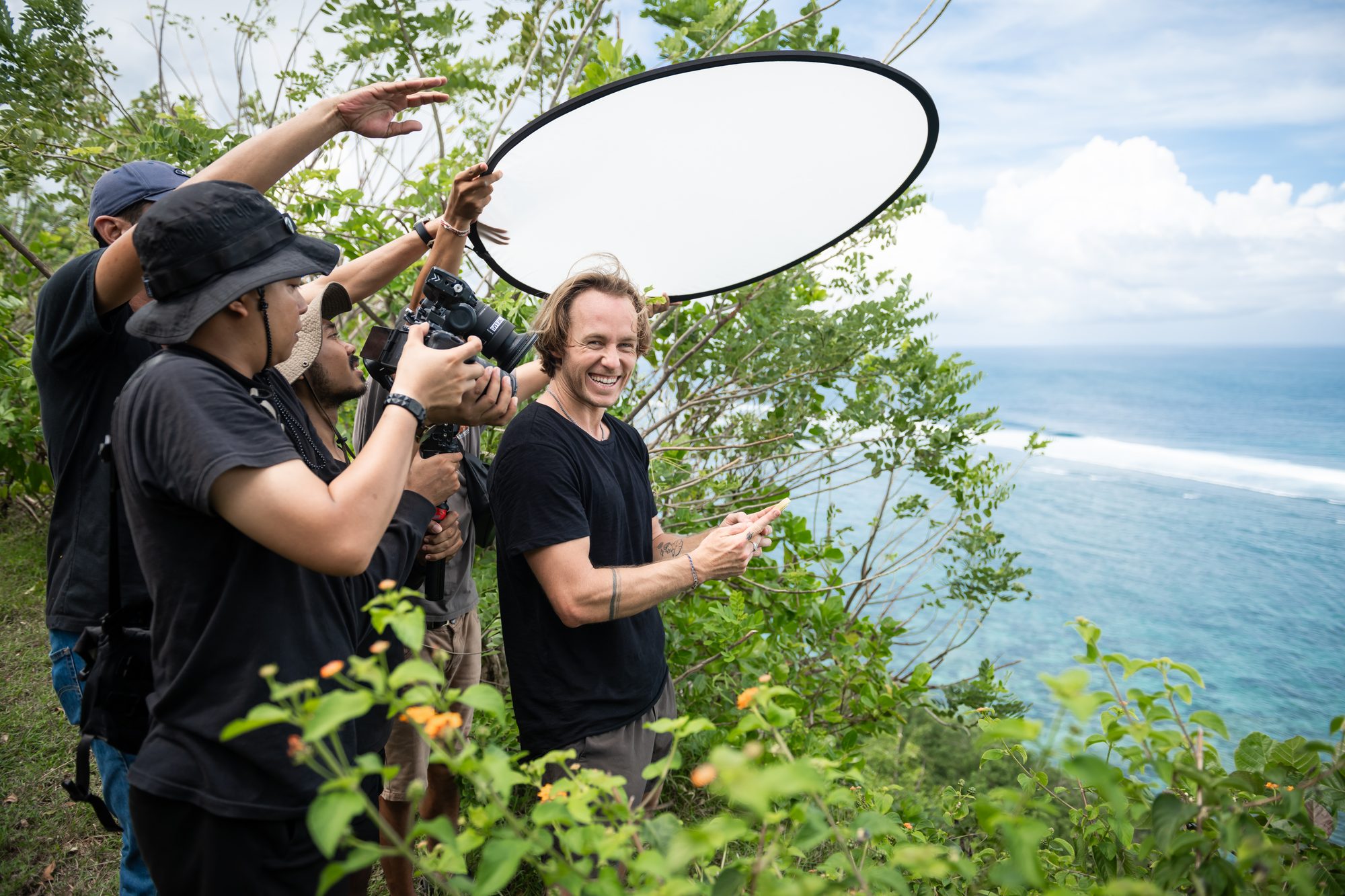 Jaro laughing on a cliffside shoot in Bali while the crew holds a reflector and camera