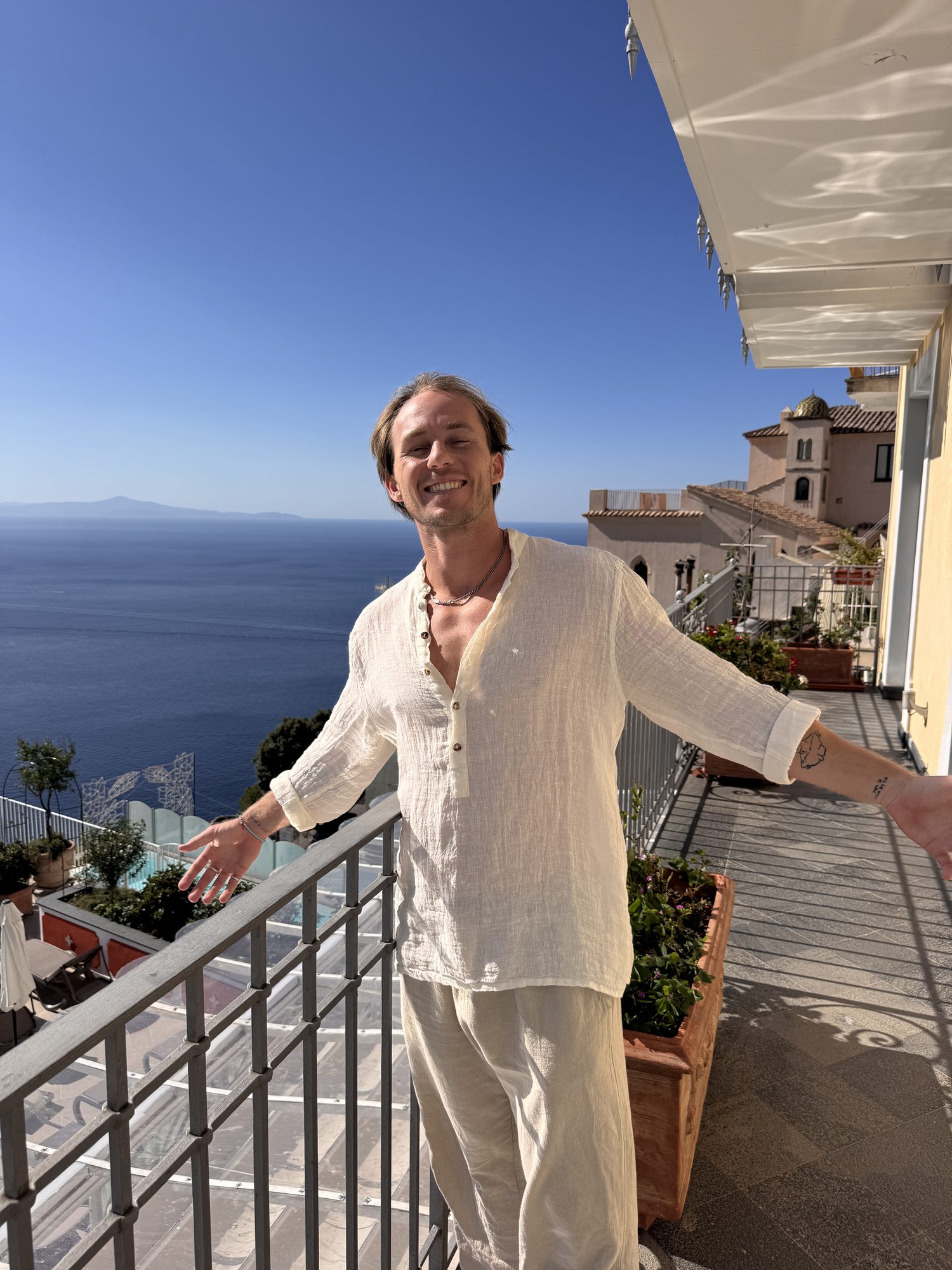 Jaro smiling with arms open on a sunny balcony above the Amalfi coast